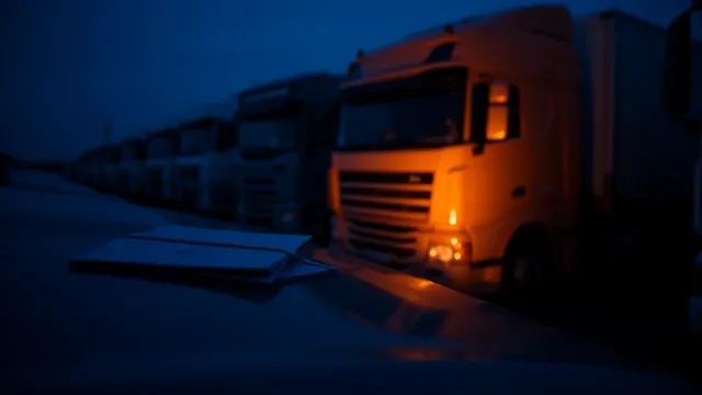 Fila de camiones aparcados en una campa logística al anochecer, con una cabina iluminada en naranja y una carpeta de documentos en primer plano